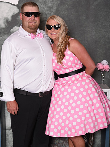 A man dressed in black pants, white shirt, pink and white bow tie, and wearing sunglasses with a woman dress in a pink dress with white spots and wearing sunglasses