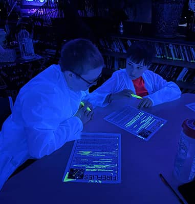 Two students in white lab coats in black lit room studying flyers about fireflies 