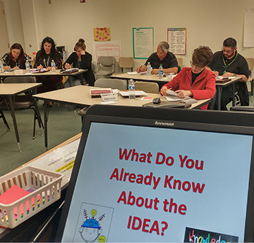 Students at desks with a sign in front that says What do you already know about the IDEA?