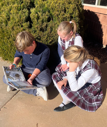 Three students looking at a book while sitting on the sidewalk