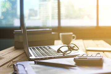 Laptop, glasses, and cup on a desk next to a window