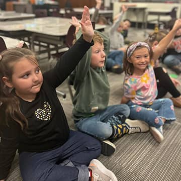 Students sitting on classroom floor with their hands raised