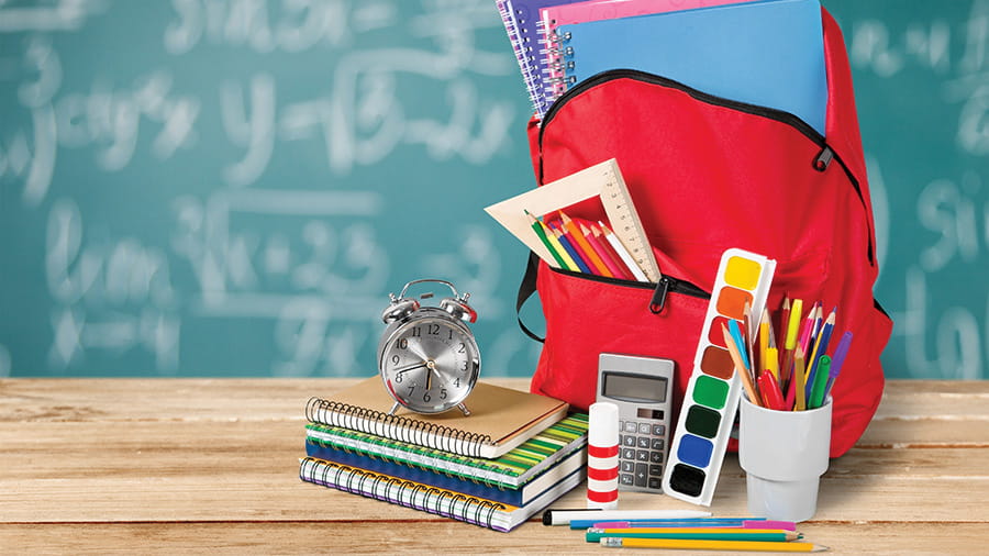 Backpack filled with notebooks and pencils, an alarm clock on a stack of notebooks, a calculator, glue stick, watercolor paints, more pencils