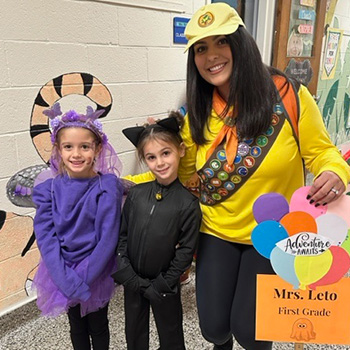 Notebook on desk in the classroom Two Students and an adult in costume holding a sign that reads Mrs. Leto First Grade