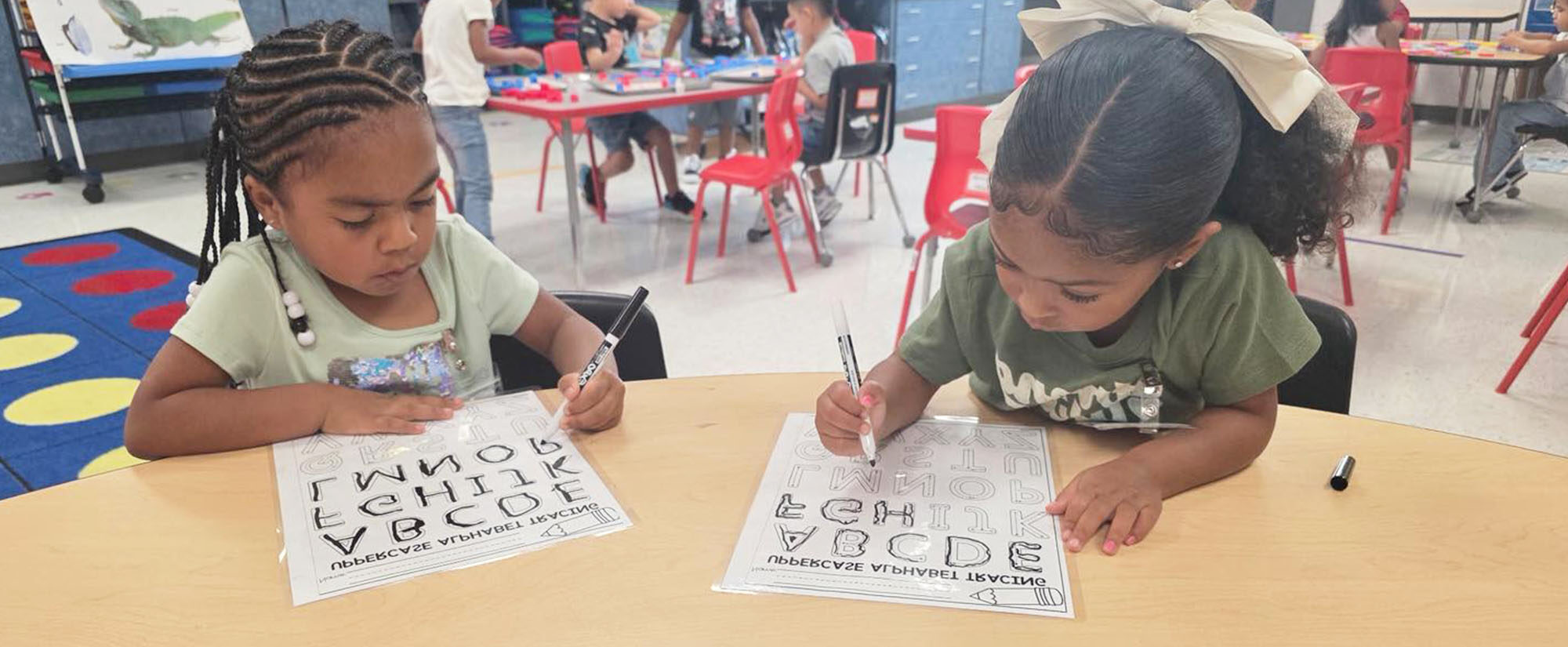Two school girls focused on an alphabet worksheet at a desk in a classroom