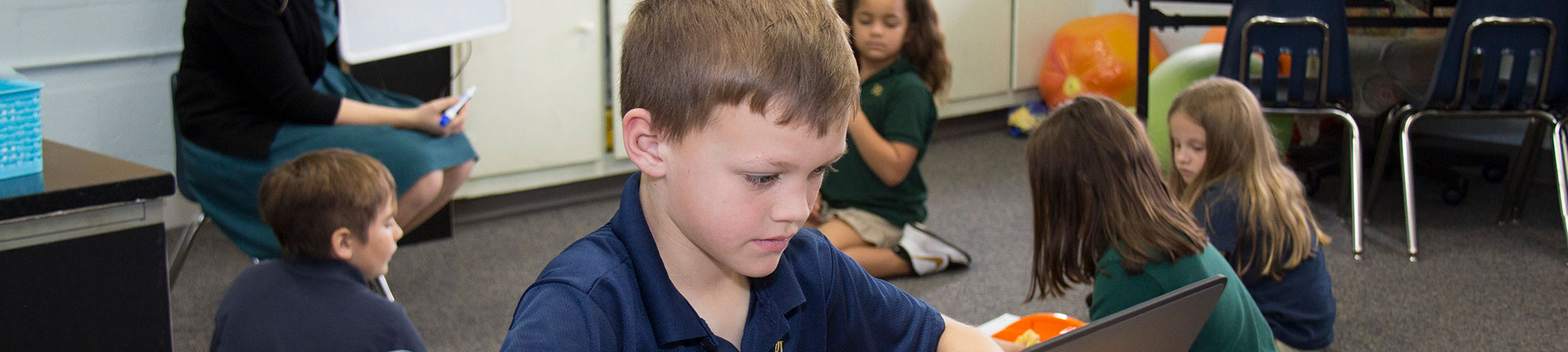 Student working on a computer with other students and a teacher in the background