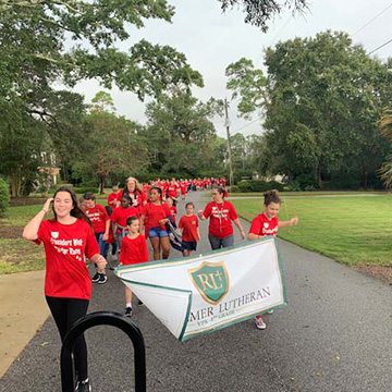 Adults and students dressed in red shirts marching with a Redeemer Lutheran banner