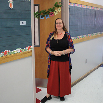 A teacher greeting people in outside a door that has a sign reading Welcome