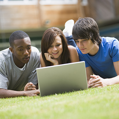 Three students laying on grass and looking at a laptop together