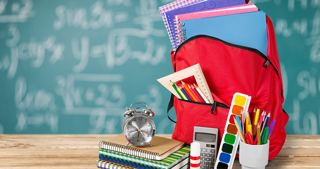 Backpack filled with notebooks, pencils, etc., on a desk with a cup of pencils, paint, glue stick, calculator, notebooks, and alarm clock