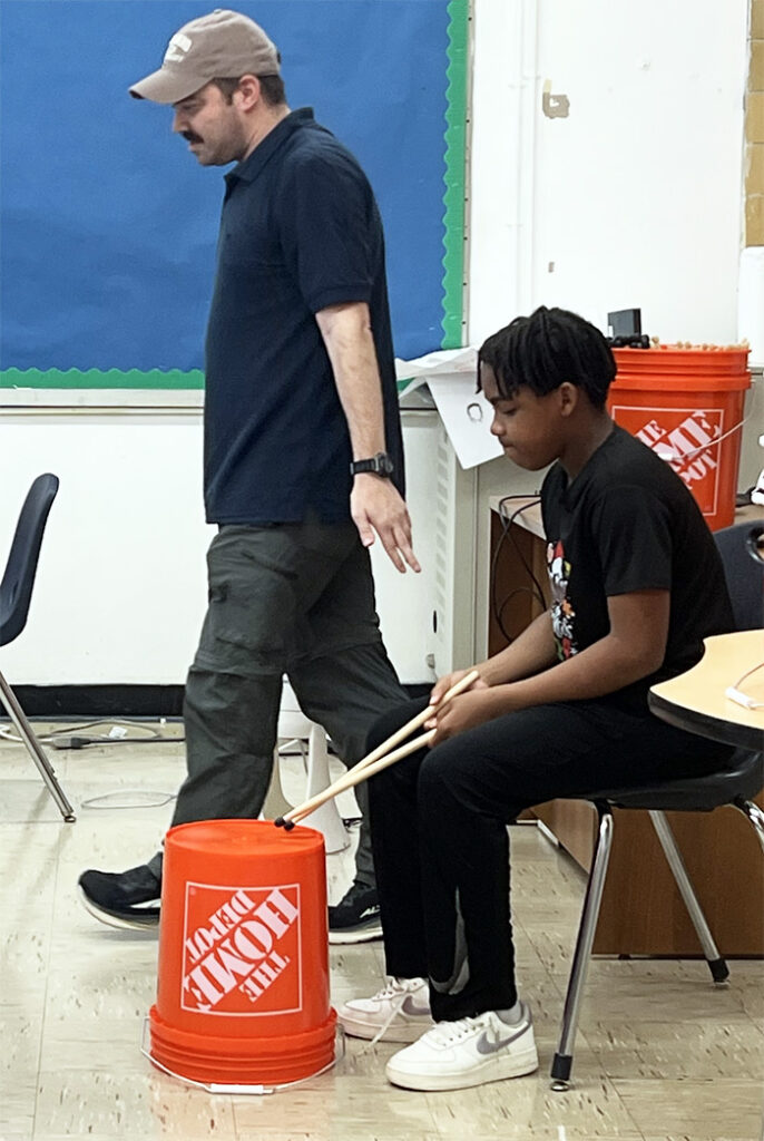 Teacher next to a student playing the drums using a Home Depot bucket