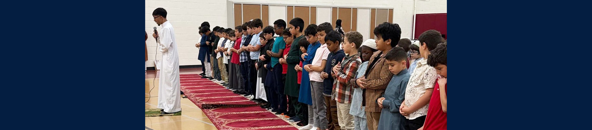 graphic-header-AboutUs Students quietly lined up in the school gym listening to a fellow student talking into a microphone