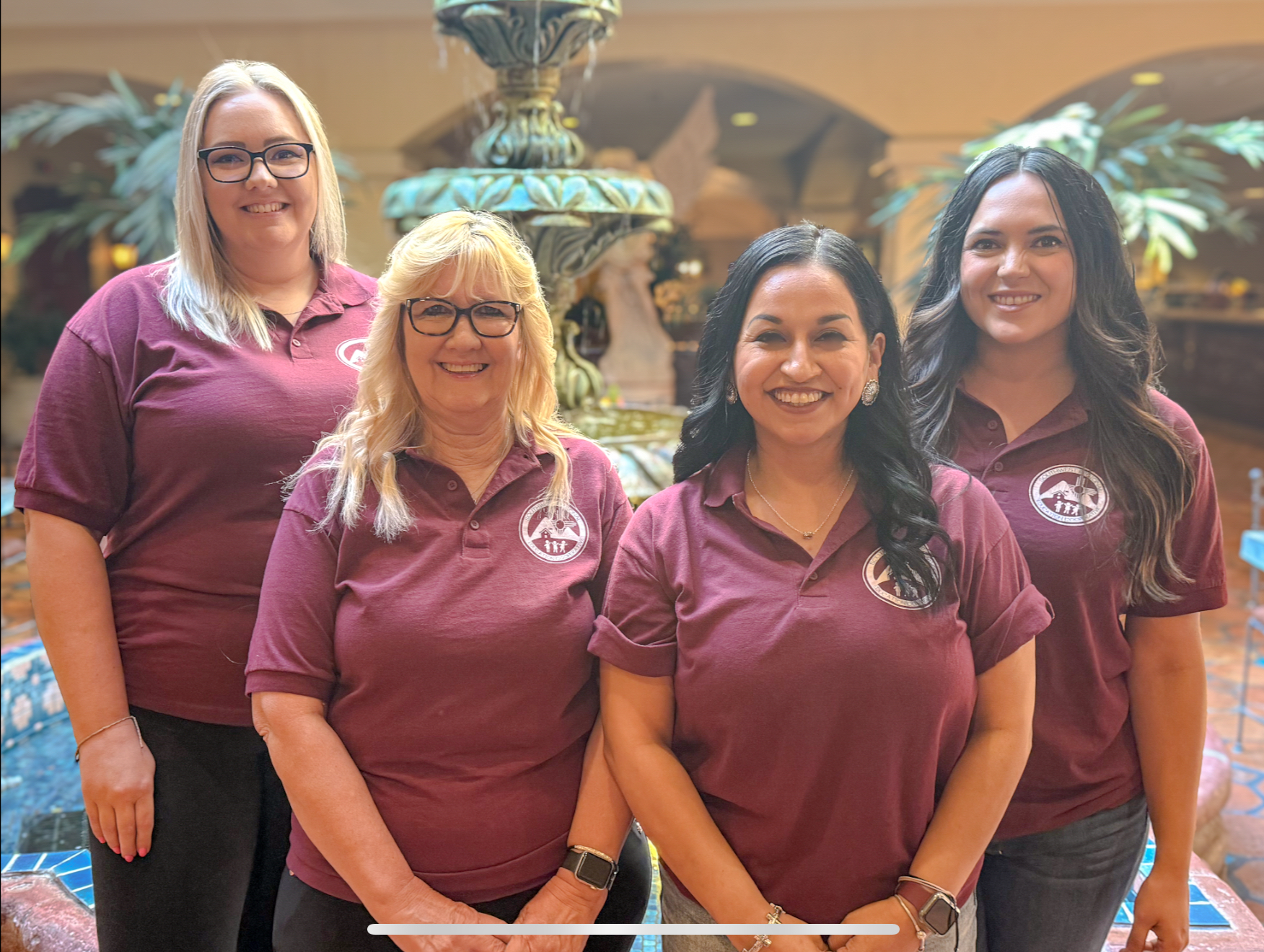A group photo of four smiling women from the SWREC Business Services team, standing in front of a decorative fountain. From left to right: Lisa Pastran, Business Services Specialist; KaLee Smyer, HR Director; Lauren Verduzco, IGA Coordinator & Chief Procurement Officer; and Erica Reyes, Finance Director. They are all wearing matching maroon polo shirts with the SWREC logo.