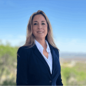 A professional headshot of Margo Martinez, a woman with long, blonde hair and a warm smile. She is wearing a dark blue blazer over a white shirt, standing in front of a blurry outdoor background with a clear blue sky.