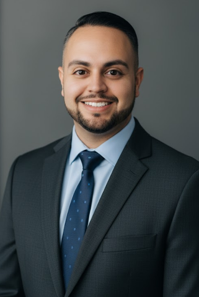 Professional headshot: A light-skinned man with dark hair and a neatly trimmed beard wearing a dark gray suit, a light blue shirt, and a navy blue patterned tie. He is smiling directly at the camera, and the background is a smooth, solid gray.
