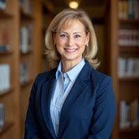 A professional headshot of Lorie Gerkey. She is a woman with shoulder-length blonde hair and a warm smile, wearing a light blue button-down shirt under a dark navy blazer. The background is a softly blurred library or office with wooden bookshelves.