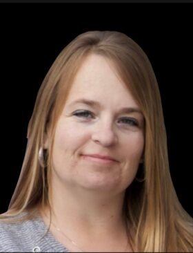 A headshot of Michelle Williams, a Caucasian woman with long light brown hair and a warm smile. She is wearing a gray sweater and large silver hoop earrings against a black background.