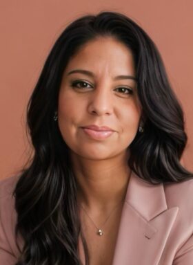 A headshot of Maria Miller, a Latina woman with long dark brown hair and brown eyes. She is wearing a light pink blazer and a silver necklace with a small gold bead. She has a subtle smile and is looking directly at the camera.