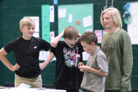 4 boys standing at a microphone looking happy and surprised