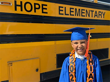 Student in a graduation cap and gown standing in front of a school bus that says Hope Elementary Student in a graduation cap and gown standing in front of a school bus that says Hope Elementary on the side