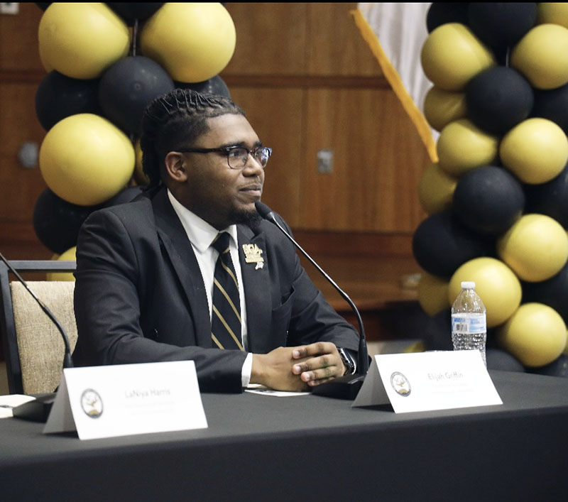 Elijah sitting at a conference table with a microphone