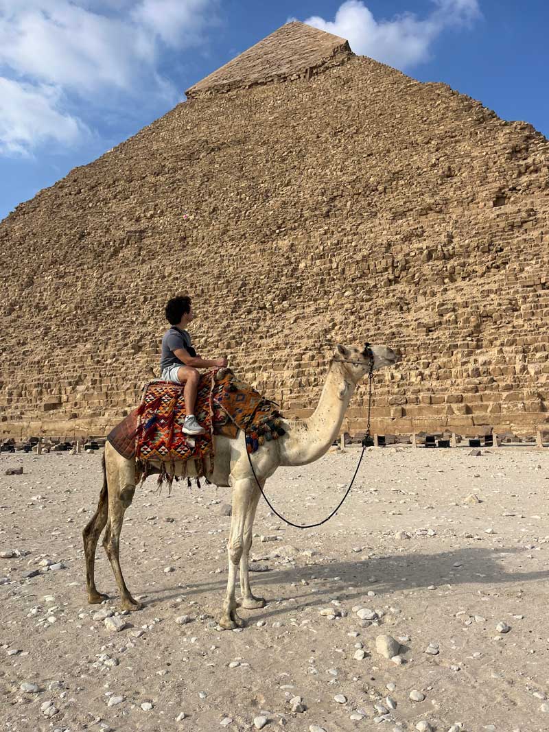 Alex riding a camel next to a pyramid